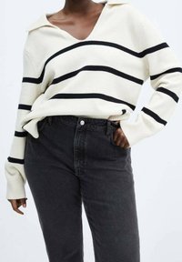 Suéter crema con rayas azul marino, escote en V y cuello, combinado con jeans negros de cintura alta. Textura suave, corte relajado. Diseño minimalista.