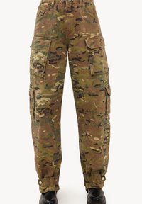 Pantalon cargo camouflage avec plusieurs poches à rabat et poignets réglables à boutons, porté avec des chaussures noires, présenté sur fond blanc.
