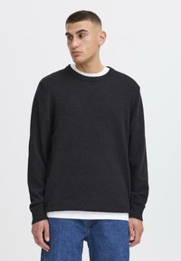 Jeune homme avec les cheveux courts, portant un pull noir sur une chemise blanche et un jean bleu, regardant légèrement vers sa gauche sur un fond uni.