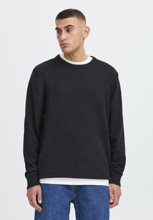 Young man with short hair wearing a black sweater over a white shirt and blue jeans, looking slightly to his left against a plain background.