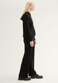 Schwarzer Hoodie mit einer Fronttasche und lässiger Passform, kombiniert mit weiten schwarzen Hosen. Beide Kleidungsstücke sind aus einem strukturierten Stoff gefertigt.