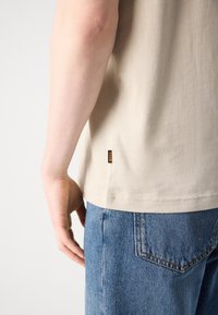 Tricou din bumbac bej cu etichetă pe lateral și mâneci scurte, combinat cu blugi din denim bleu deschis, având un buzunar și cusături în contrast.