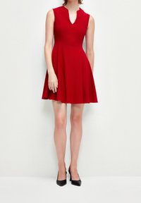 Czerwona sukienka bez rękawów z dopasowanym górą i rozkloszowanym dołem, z dekoltem w kształcie litery V. Materiał wydaje się gładki i lekki. Noszona z czarnymi szpilkami.