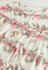Robe en tissu blanc ornée d'un motif floral avec des nœuds rouges, des branches de pin et des baies. Comprend des détails froncés et un cordon de serrage.