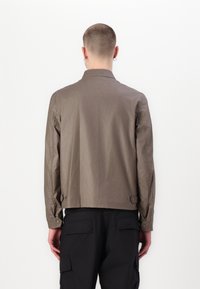 Světlohnědá lehká bunda s přímo střiženým, strukturovaným límcem a manžetami na knoflíky, v kombinaci s černými nákladními kalhotami.