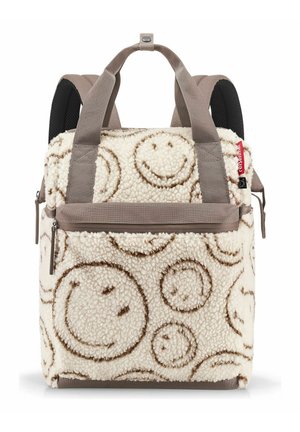 Zaino color crema con motivi di faccine sorridenti marroni, spallacci taupe, tasca frontale con zip e etichetta rossa del marchio sul lato.