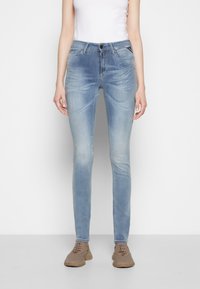 Donna che indossa jeans skinny sbiaditi di un blu chiaro, top bianco senza maniche e sneakers in rete beige, in piedi contro uno sfondo bianco semplice.