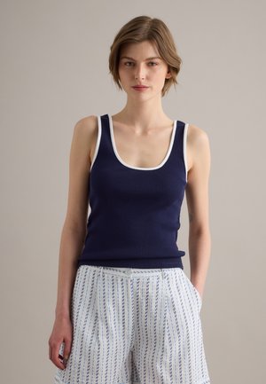 Femme portant un débardeur bleu marine avec bordure blanche et un short blanc à motifs avec des rayures verticales bleues, debout sur un fond uni.
