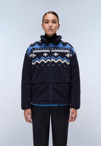 Giacca in pile blu navy con motivi geometrici crema e blu, colletto alto, chiusura con zip e tasche laterali, indossata sopra un dolcevita nero.