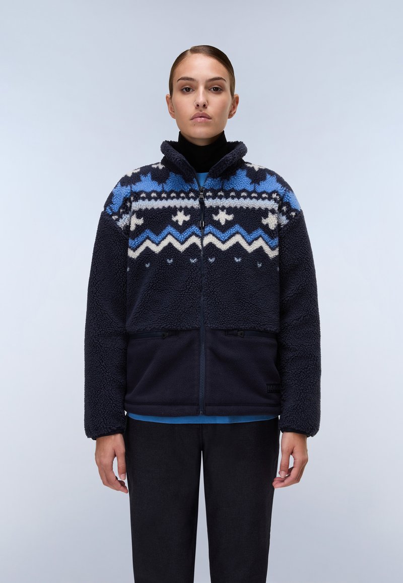 Giacca in pile blu navy con motivi geometrici crema e blu, colletto alto, chiusura con zip e tasche laterali, indossata sopra un dolcevita nero.