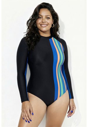 Femme aux longs cheveux foncés portant un maillot de bain noir à manches longues avec des rayures verticales bleues, orange et vertes, souriante à la caméra.