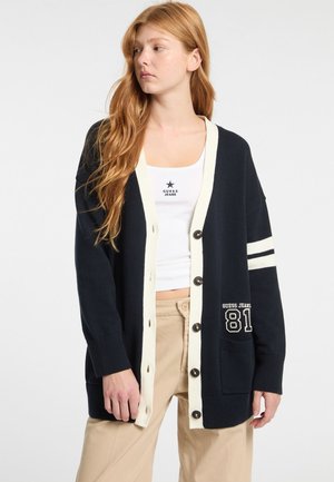 Cardigan blu navy con bordi cream, dotato di grandi bottoni, due tasche frontali e strisce bianche sulle maniche. Materiale in maglia di cotone.