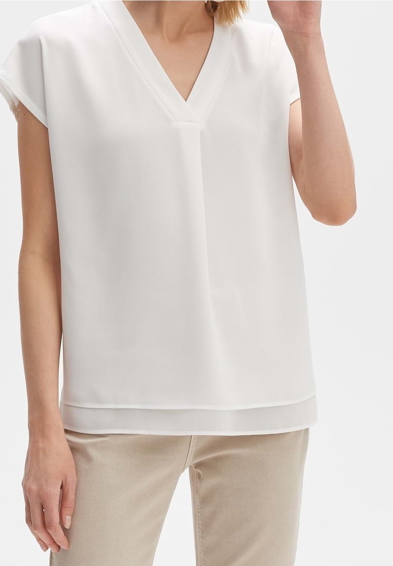 Blouse blanche à manches courtes avec un décolleté en V, fabriquée en tissu lisse. Présente un ourlet superposé pour plus de détail. Associée à un pantalon beige.
