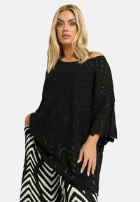 Mustikka, neulottu oversized villapaita, jossa on avoin kudontamalli ja kellohihat, yhdistettynä mustavalkoisiin sahalaitakuosisiiin housuihin.