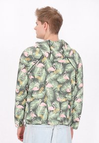 Giacca con cappuccio in tessuto leggero, con sfondo verde e fenicotteri rosa accompagnati da foglie tropicali. Dettagli con polsini e orlo elasticizzati.