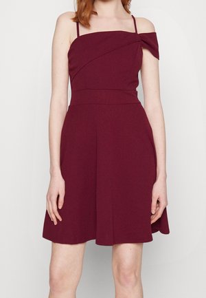 Kvinde iført en ærmeløs, skulderfri, burgunder knælængde kjole med tynde stropper mod en ensfarvet baggrund.