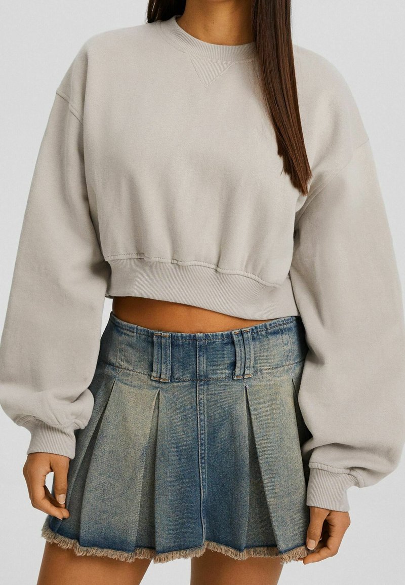 Sudadera beige recortada con mangas abullonadas, combinada con una falda de mezclilla plisada con un dobladillo deshilachado y un degradado de azul claro a oscuro.