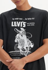 Czarny bawełniany t-shirt z vintage'owym białym nadrukiem przedstawiającym kowboja na koniu, z napisem "LEVI'S THE ORIGINAL BLUE JEANS". Krój z okrągłym dekoltem.