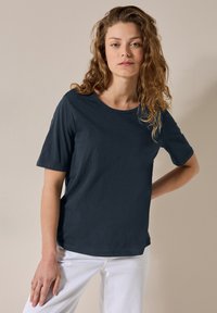 Tmavě modré tričko s krátkým rukávem, kulatým výstřihem, vyrobené z měkké tkaniny. V kombinaci s bílými kalhotami, vystavené na neutrálním pozadí.