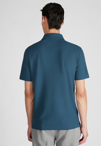 Polo verde petrolio scuro con colletto, maniche corte e orlo diritto. Texture liscia, dettagli minimi, indossata con pantaloni grigi chiari.