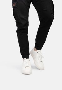 Sneakers bianche con lacci, dalla texture liscia, suole in gomma e design minimalista, abbinate a pantaloni cargo neri con polsini elasticizzati.