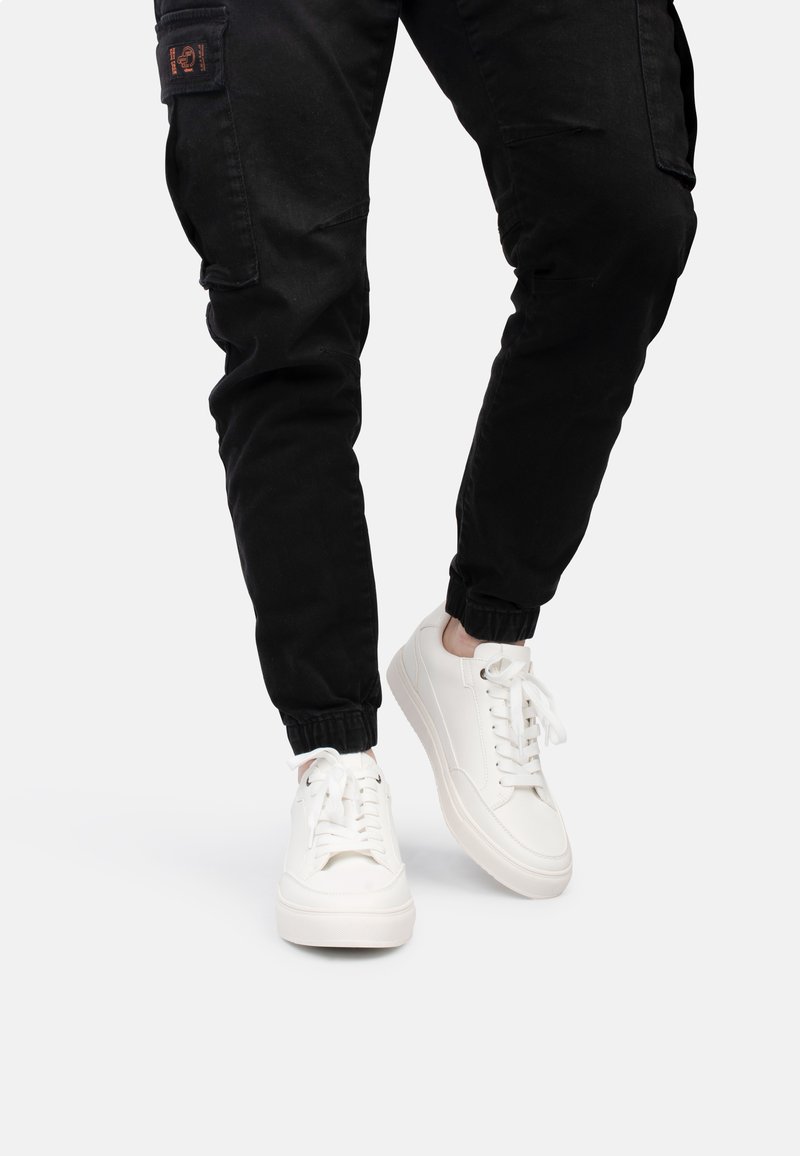 Sneakers bianche con lacci, dalla texture liscia, suole in gomma e design minimalista, abbinate a pantaloni cargo neri con polsini elasticizzati.
