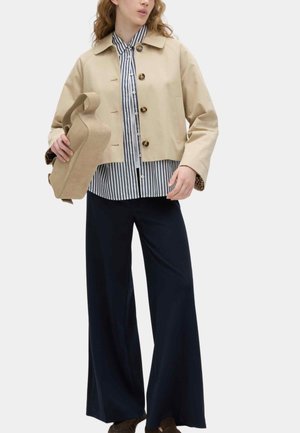 Kvinde iført beige cropped jakke over stribet skjorte, vidt ben marineblå bukser, holder en beige struktureret håndtaske mod hvid baggrund.