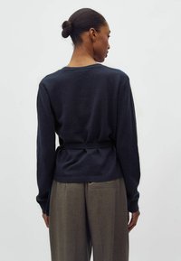 Sieviete ar matiem sasietiem bulciņā, valkā tumši zilu džemperi ar jostu un brīvas pelēkas bikses, stāvot pret vienkāršu gaišu fonu.