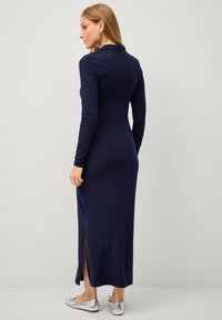 Navy-blaues, geripptes Strickkleid mit langen Ärmeln und einem Schlitz vorne, das eine figurbetonte Passform und einen klassischen Kragen hat. Dazu werden silberne Ballerinas getragen.