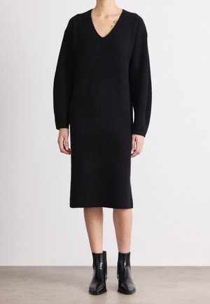 Kvinde iført en lang sort strikket kjole med V-hals og lange ærmer, kombineret med sorte ankelstøvler, stående på en grå gulv.