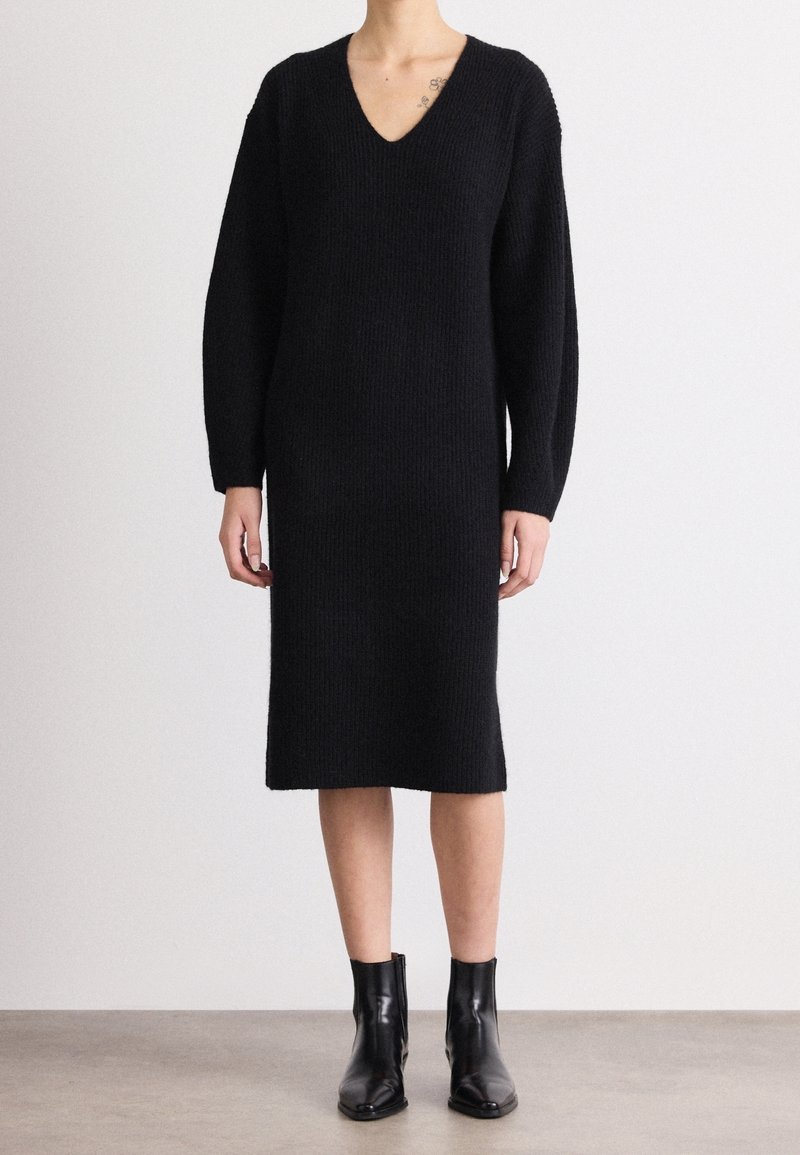 Femme portant une longue robe en tricot noire avec un col en V et des manches longues, associée à des bottines noires, debout sur un sol gris.