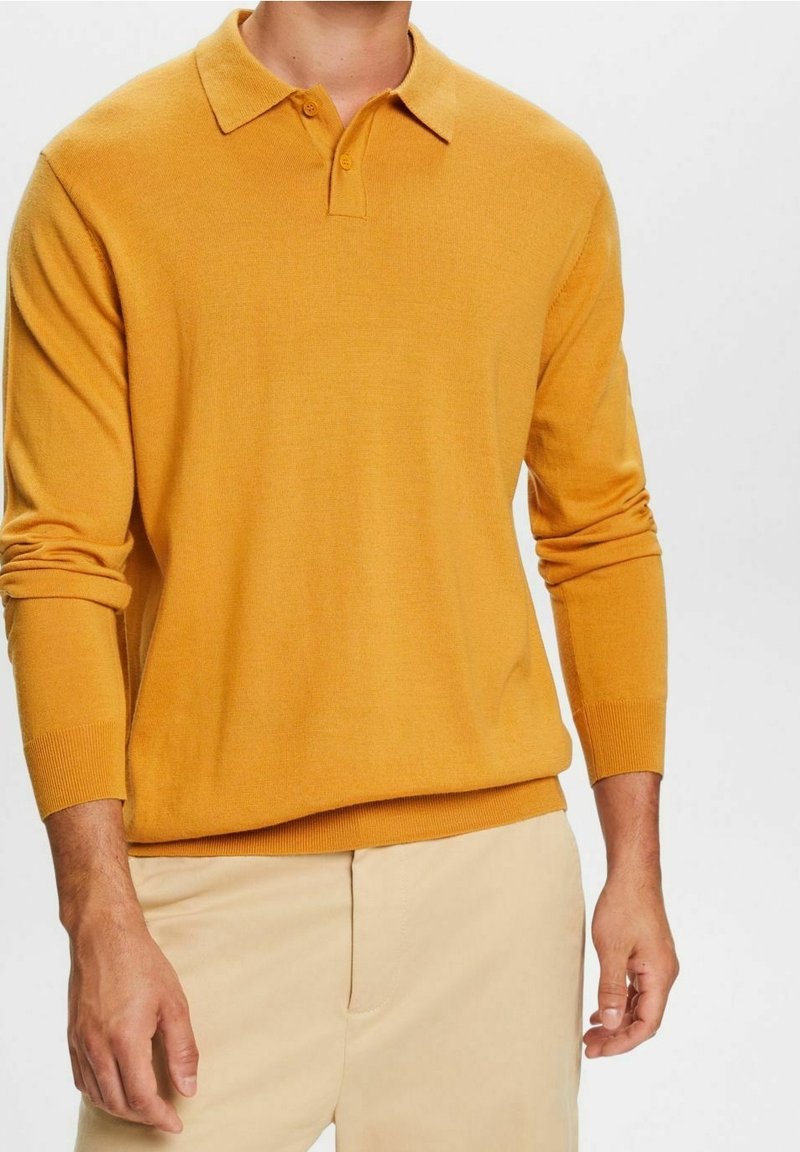 Langärmliges Poloshirt in senfgelb, gestrickte Textur, Kragen mit zwei Knöpfen, gerippte Bündchen und Saum, kombiniert mit hellbeigen Hosen.