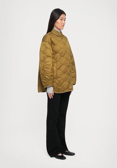 Olīvzaļš quiltotais mētelis ar apaļu apkakli, ar pogu aizdari un lielu piegriezumu. Valkāts virs gaiši pelēka krekla un melnām biksēm.