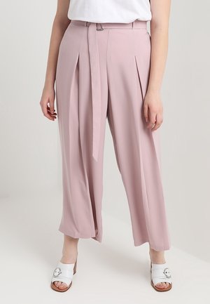 Femme portant un pantalon rose taille haute à jambes larges avec une ceinture assortie en tissu, un haut sans manches blanc et des sandales blanches à talons.
