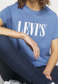 Personne assise avec un genou relevé, portant un t-shirt bleu Levi's, un jean bleu foncé et des baskets blanches, cadrée au niveau du cou et du visage.