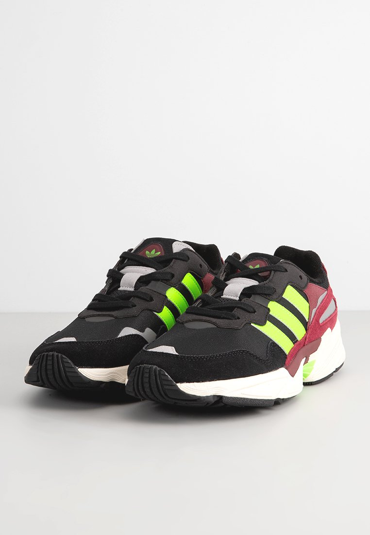 Svarta träningsskor med gröna ränder, röda accenter och en kombination av nät och mocka material. Har en vadderad krage och gummisula.