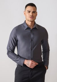 Camicia grigia con bottoni, maniche lunghe, tessuto morbido, colletto classico e bottoni neri, abbinata a pantaloni scuri. Design semplice e su misura.