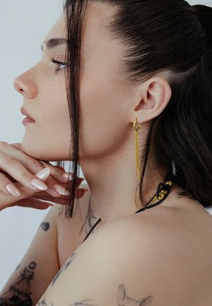 Frau mit dunklem Haar, die einen goldenen, hängenden Ohrring trägt, ihr Kinn auf die Hände stützt und Tätowierungen am Oberarm und der Schulter zeigt.