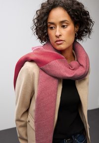 Donna con capelli ricci che indossa orecchini a cerchio dorati, una giacca beige, una maglietta nera, jeans blu e una grande sciarpa rosa avvolta attorno alle spalle.