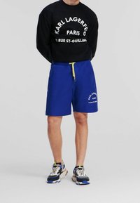 Camiseta negra de manga larga con texto blanco y pantalones cortos azules con cordón amarillo. Zapatillas deportivas con detalles multicolores.