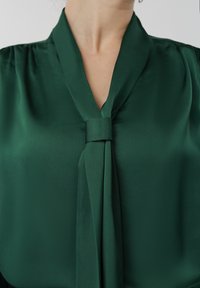 Femme portant un chemisier vert foncé à col en V avec un détail noué en tissu au col, cadrée du cou jusqu'au milieu du torse.