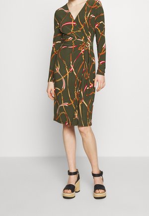 Femme portant une robe portefeuille olive mi-longue avec un imprimé chaîne multicolore et des sandales plateformes noires à brides autour de la cheville.