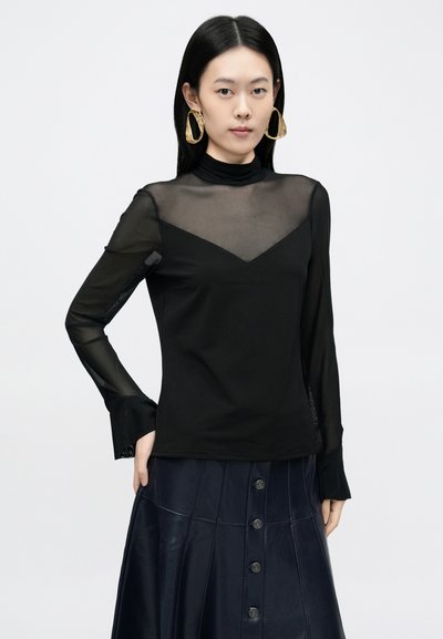 Fekete hosszú ujjú felső hálós réteggel és magasított nyakkal. A mellrészen cikk-cakk mintával, valamint bővülő mandzsettákkal rendelkezik.