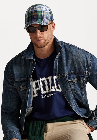 Farmerdzseki, tengerészkék póló "POLO" felirattal, zöld és kék árnyalatú kockás sapka, fekete napszemüveg, világos bézs nadrágban ül.