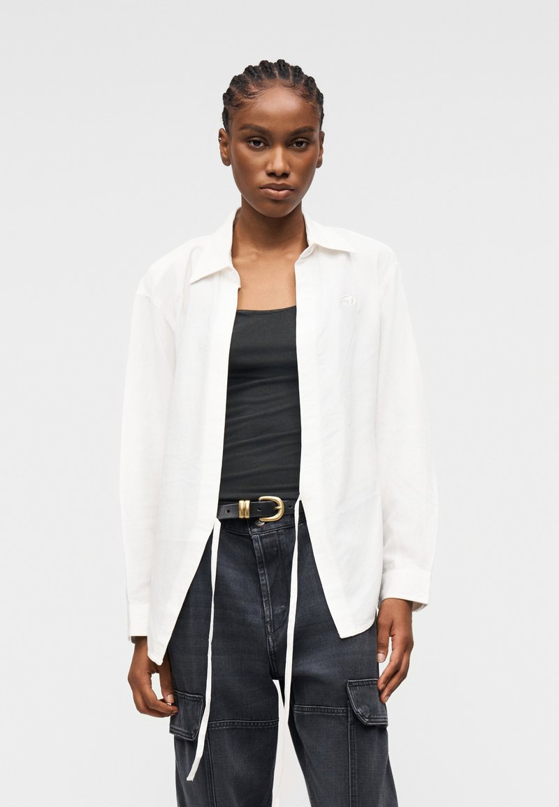 Γυναίκα με πλεξουδάκια που φοράει λευκό ανοιχτό πουκάμισο πάνω από μαύρο τοπ, μαύρη ζώνη και σκούρο παντελόνι cargo, κοιτάζοντας ευθεία με ουδέτερη έκφραση.