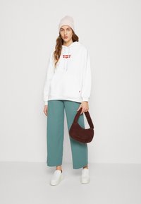 Biela mikina s kapucňou a červeným logom, kombinovaná so zelenomodrými nohavicami s voľným strihom, bielymi slip-on topánkami a hnedou textúrovanou kabelkou. Na hlave má ružovú čiapku.