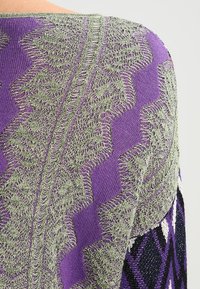 Pullover lavorato a maglia con un design geometrico viola e verde, tessuto testurizzato e un colletto leggermente aperto con cuciture dettagliate.