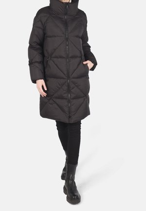 Person iført en lang sort quiltet dunjakke med lynlås foran, sorte leggings og sorte ankelstøvler, gående med den ene hånd i lommen.