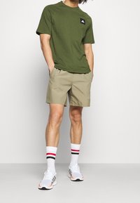 Oliwkowa koszulka z czarnym logo, khaki szorty i białe sneakersy. Białe skarpetki w czerwone i czarne paski. Casualowy strój sportowy.