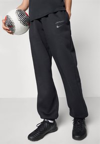 Svarte joggebukser med løs passform, med en liten grå Nike-logo og teksten "SPURS". En strukturert hvit og svart fotball holdes.